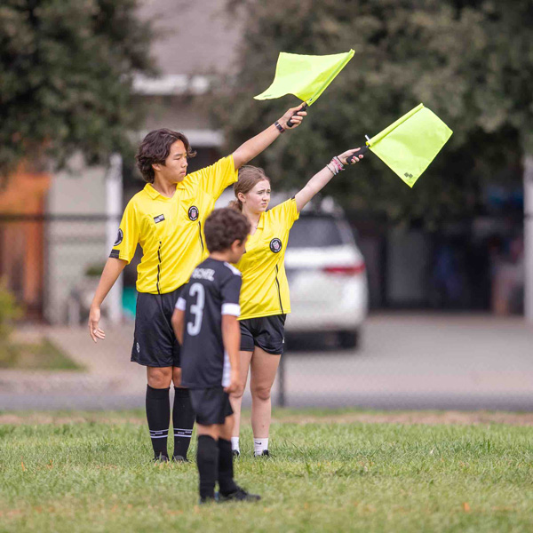 Two teenage soccer referees both pointing their flag while officiating a youth recreational soccer league game.