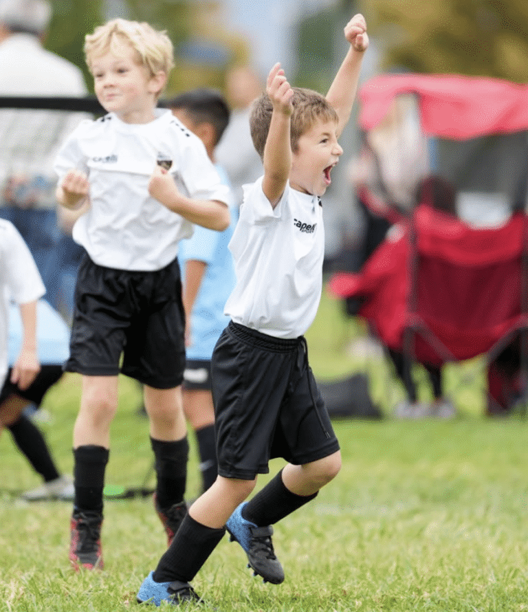 Two young soccer players celebrating on a field, with red tents and spectators in the background.