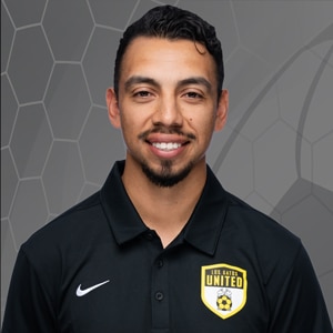 Portrait of Coach Jesus Rodriguez, a smiling man with short dark hair and goatee wearing a black polo shirt featuring a United soccer club crest against a gray geometric background.