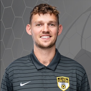 Portrait of a smiling male soccer player wearing a dark striped polo with a yellow United crest on the chest and a Nike swoosh.