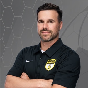 Male soccer coach with short dark hair and beard, wearing a black polo with a yellow team crest, arms crossed and a neutral smile.