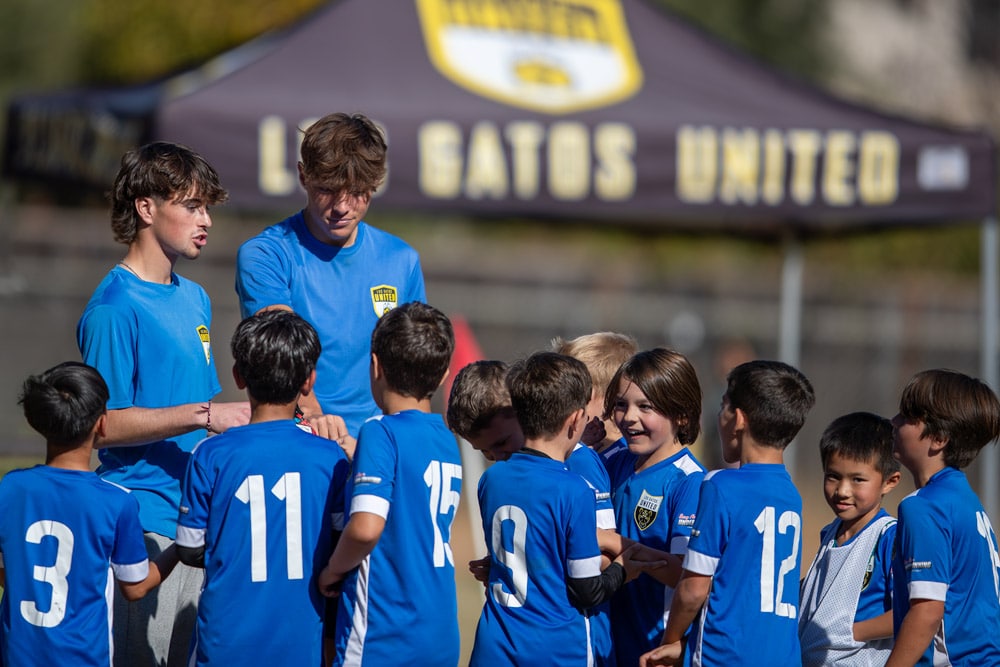 Young soccer players in blue uniforms gather around two coaches on a field, under a tent that reads 'UNITED' in the background.